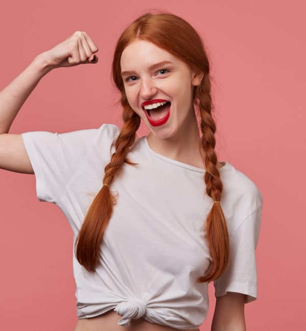 Indoor portrait of young ginger female with freckles posing over pink background showing her biceps and smiles broadly Nutricionista SOP - ¡Reserva ya tu consulta online!