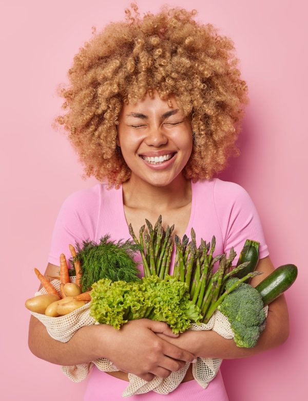 Natural food concept. Joyful woman with curly hair keeps eyes closed smiles toothily poses with freshly picked potatoes carrot asparagus and lettuce isolated over pink background. Eco products Alimentos saludables para la endometriosis