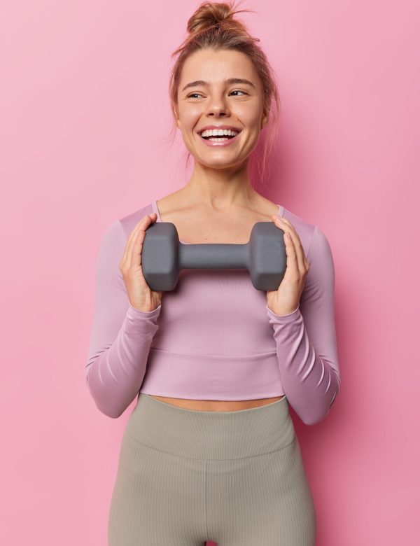 Vertical shot of healthy fit sportswoman has regular training in gym holds dummbell smiles gladfully leads active lifestyle dressed in tracksuit isolated over pink background. People and sport Vertical shot of healthy fit sportswoman has regular training in gym holds dummbell smiles gladfully leads active lifestyle dressed in tracksuit isolated over pink background. People and sport